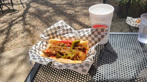 Sausage and pepper sub with red sauce and caramelized onions in a checkered paper-lined basket with a drink on a metal table.