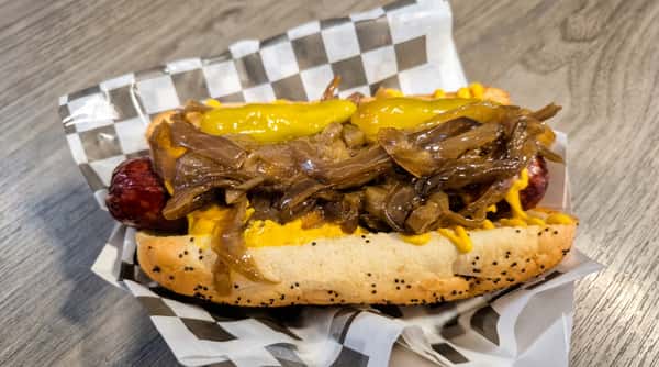 Hot dog in a poppy seed bun with yellow mustard, caramelized onions, and pale green peppers.
