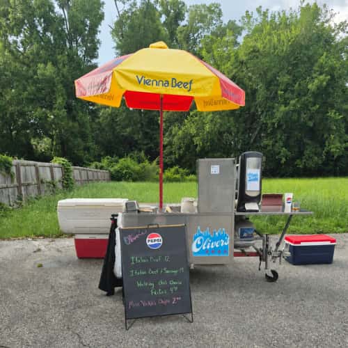 A "Vienna Beef" hot dog cart with a red and yellow umbrella, facing a grassy, tree-lined background.