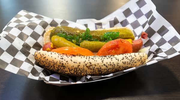 Hot dog in a poppy seed bun with relish, pickles, tomato slices, and mustard in a checkered paper tray.