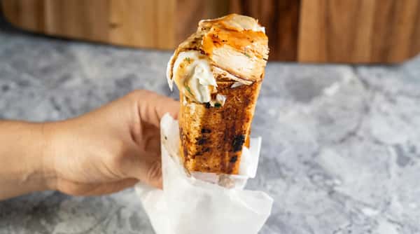 Hand holding a grilled sandwich filled with white meat chicken and creamy sauce, wrapped in paper.