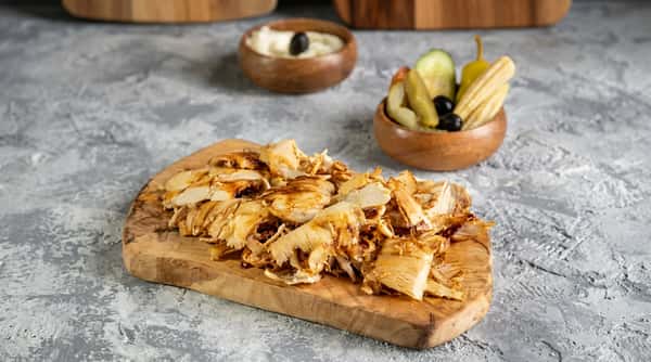 Shawarma chicken on a wooden board, with sides of pickled vegetables and a white dipping sauce with an olive.