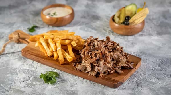 Shawarma platter with sliced meat, french fries, a side of pickles and olives, and a dipping sauce.
