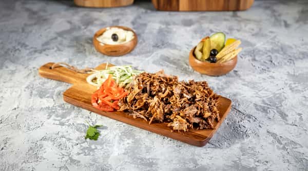 Shawarma meat on a wooden board with sliced onions, tomatoes, and parsley. Side bowls of pickles and creme.