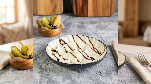 Creamy white dip drizzled with dark sauce in a black bowl, with pickles and olives in background.