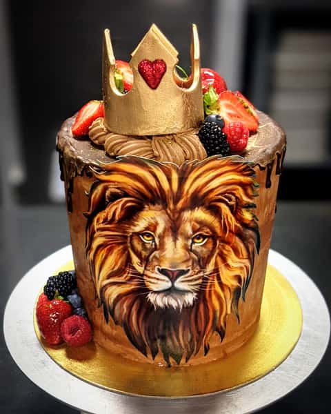 Chocolate drip cake with realistic lion portrait, fresh berries, and gold crown topper on a decorative celebration cake.