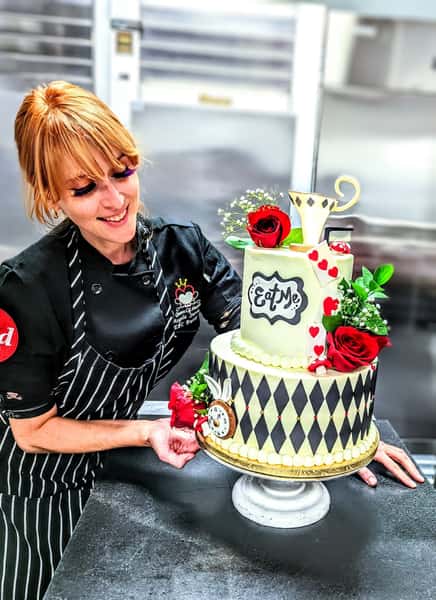 Pastry chef smiling beside a whimsical Alice in Wonderland themed cake decorated with roses, hearts, teacup, and "Eat Me" sign.