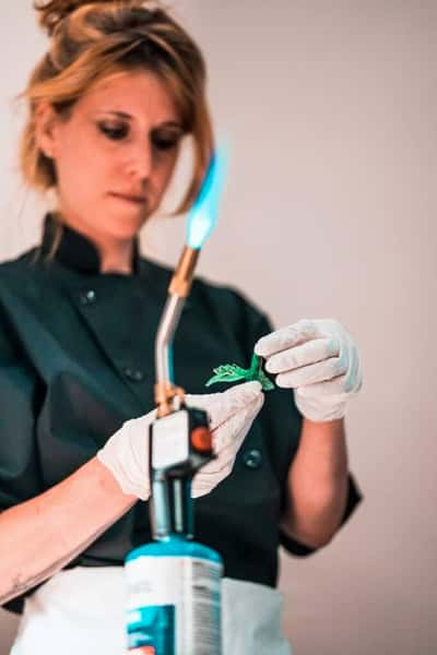 Chef using a blowtorch to caramelize a garnish while wearing gloves and a black chef coat in a kitchen setting.