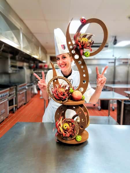 Smiling pastry chef in a professional kitchen presenting an elaborate chocolate showpiece sculpture while making peace signs.
