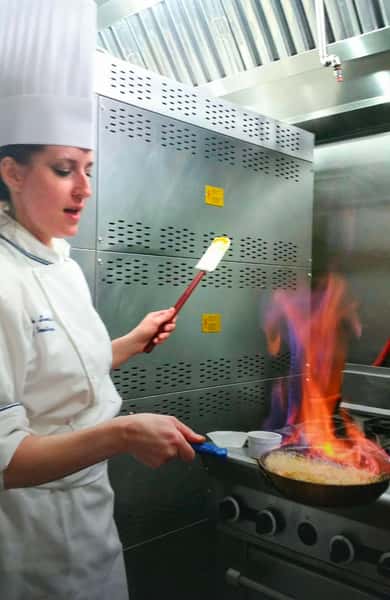 Chef in white uniform flambéing a dish in a pan over a gas stove, flames rising in a professional stainless steel kitchen.