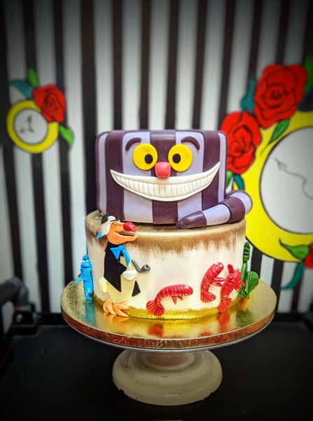 Whimsical Cheshire Cat themed cake with striped cat topper, cartoon characters, and colorful fondant decorations.