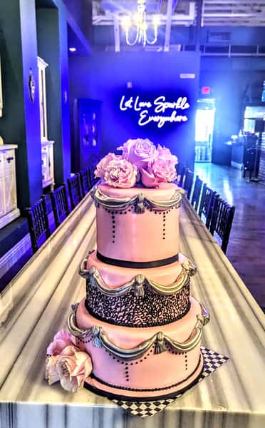 Three-tier pink and gold decorative cake with large sugar roses displayed on a marble bar beneath a glowing neon sign.