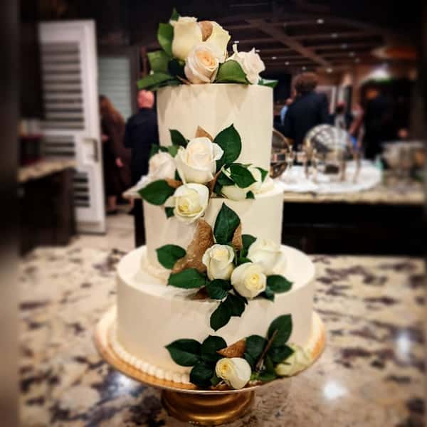 Three-tier white wedding cake with ivory roses, green leaves, and gold accents displayed on a stand at an elegant reception.