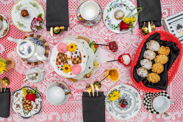 Overhead view of a whimsical afternoon tea spread featuring scones, tea sandwiches, cookies, and floral china on a lacy red tablecloth.