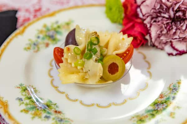 Bowtie pasta salad with artichoke, cherry tomato, green olives, and scallions, served in a small dish on a floral tea plate.