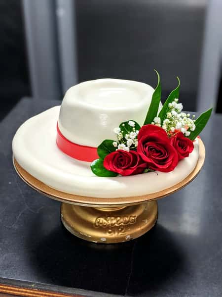 White fondant hat-shaped cake with red ribbon band and fresh red roses displayed on a gold cake stand.