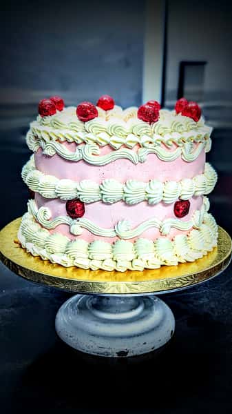 Round pink buttercream cake with ornate piped frosting swirls and red cherries displayed on a gold cake stand.