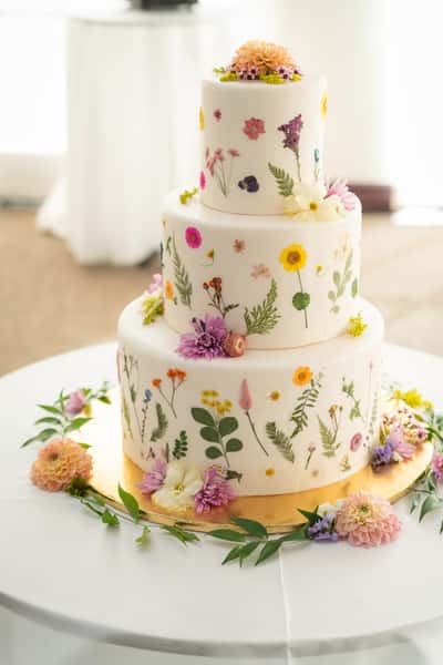 Three-tier white cake decorated with pressed edible flowers and fresh blossoms, creating a delicate botanical garden design.