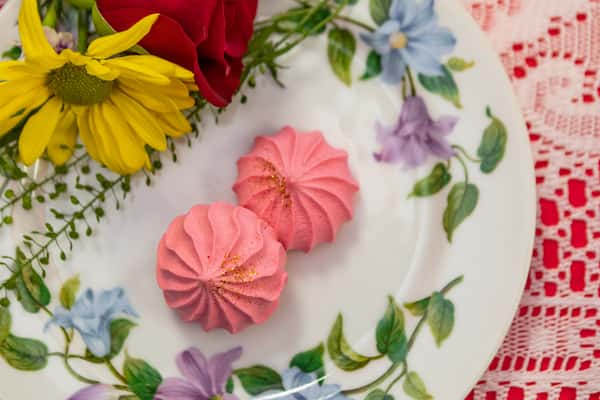 Two pink meringue cookies with a touch of edible glitter, served on a floral-patterned plate with fresh daisy and rose garnish.