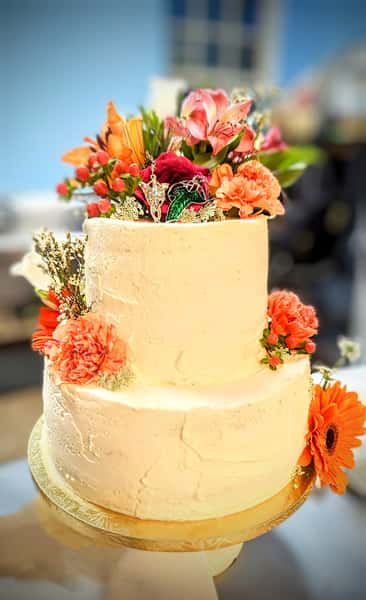 Two-tier buttercream cake decorated with vibrant fresh flowers including lilies, roses, and gerbera daisies in warm orange and pink tones.