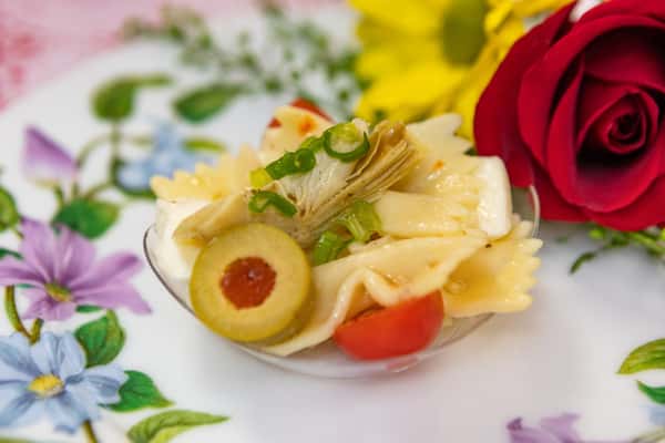 Bowtie pasta salad with artichoke, cherry tomato, green olives, and scallions, served in a small dish on a floral tea plate.