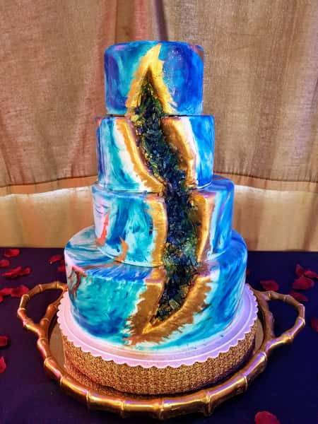 Four-tier geode cake with blue marble icing, gold accents, and a cascading crystal sugar center displayed on a gold tray.