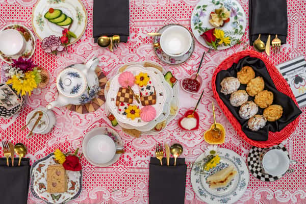 Overhead view of a whimsical afternoon tea spread featuring scones, tea sandwiches, cookies, and floral china on a lacy red tablecloth.