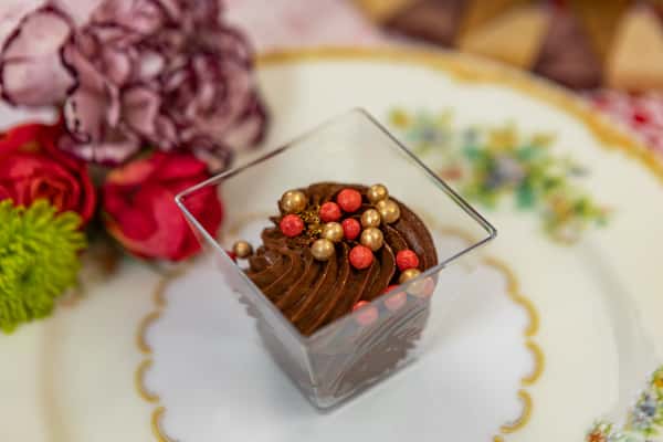 Rich chocolate mousse in a clear cup, topped with gold and red edible pearls, presented on an elegant floral porcelain plate.
