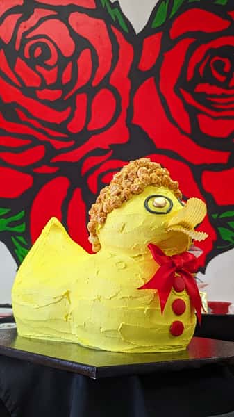 Yellow duck-shaped novelty cake with red bow tie and chips in its beak, set against a bold red rose backdrop.