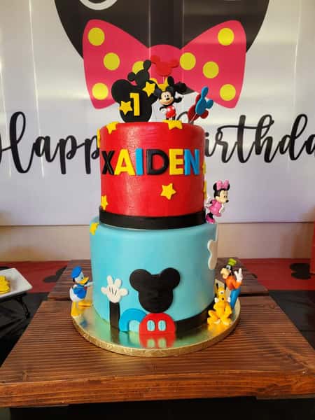 Two-tier Mickey Mouse themed first birthday cake with colorful stars and Disney characters, personalized with the name Kaiden.
