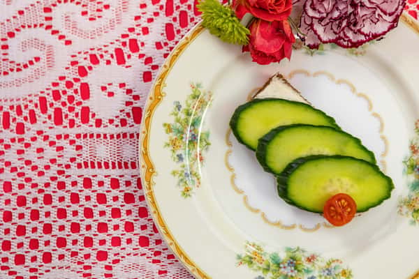 Cucumber tea sandwich on rye bread with creamy spread and cherry tomato garnish, served on ornate vintage china with floral trim.