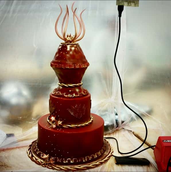 Three-tier red and gold decorative cake with ornate details and sculpted topper resembling elegant flame shaped sugar art.