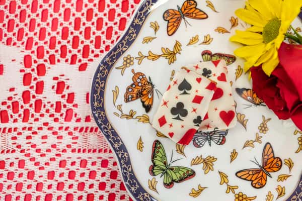 White chocolate fudge decorated with playing card suit symbols, served on a butterfly-patterned plate with a rose and daisy garnish.