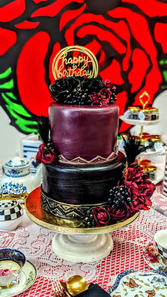 Elegant two-tier burgundy and black birthday cake with dark roses, berries, and gold Happy Birthday topper on display.