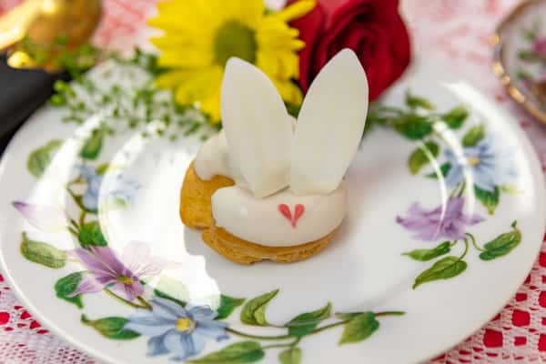 Whimsical bunny-shaped cream puff topped with white icing and fondant ears, served on a floral vintage plate with fresh flowers.