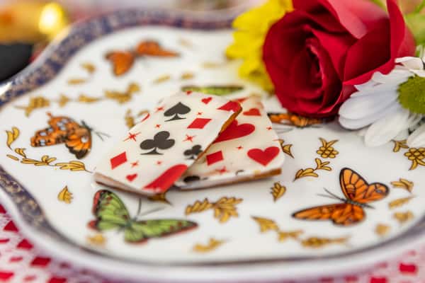 Royal icing-topped cookies with playing card suit designs, served on a butterfly porcelain plate with rose and daisy floral accents.