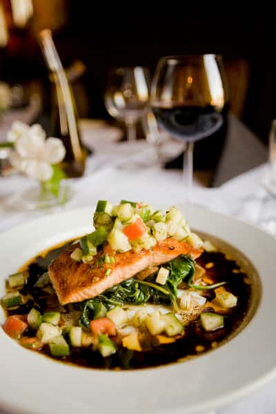 Grilled salmon fillet topped with diced vegetables on sautéed greens with sauce, served in an upscale dining setting.