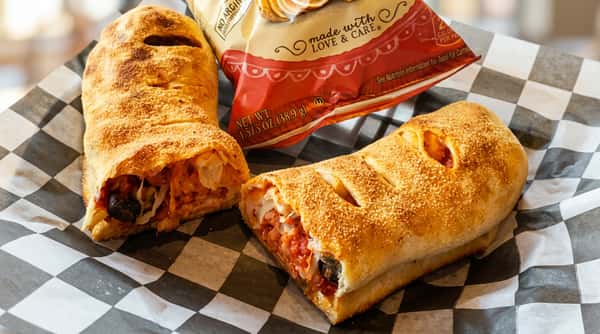 Two baked calzones, one cut open to show red sauce, meat, and cheese, on checkerboard paper with a snack bag.
