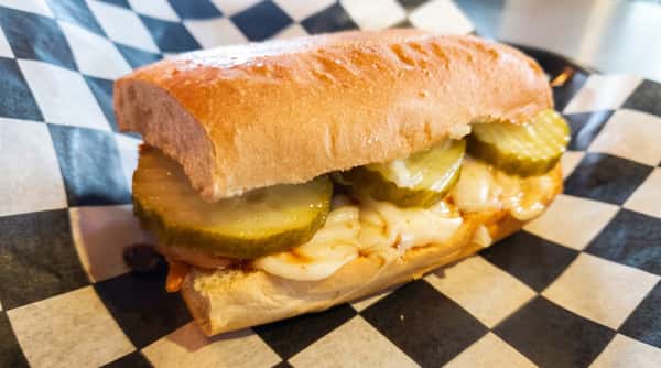 Submarine sandwich with melted white cheese, pickle slices, and reddish sauce on a checkered paper liner.