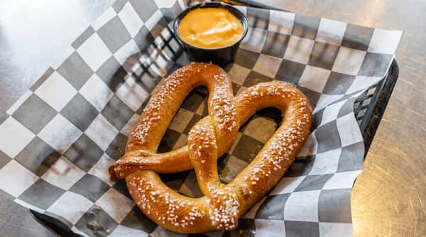 Soft pretzel with coarse salt and a side of orange cheese dip in a black cup, on checkered paper.