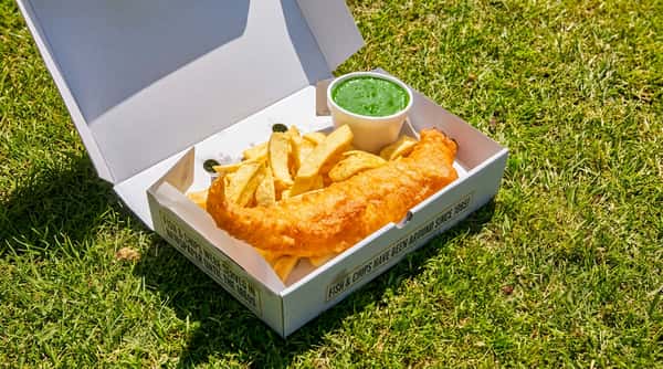 Battered fish and chunky chips served in a takeaway box with mushy peas outdoors.
