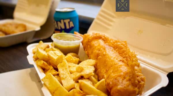 Golden battered fish with chunky chips, lemon and sauce in a takeaway box.