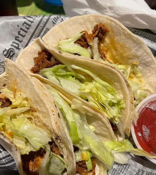 Three soft shell tacos filled with shredded meat and lettuce, served with a side of salsa.
