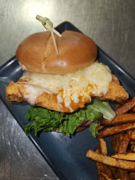 Crispy chicken sandwich topped with melted cheese and hot sauce on a toasted bun, served with golden brown fries.