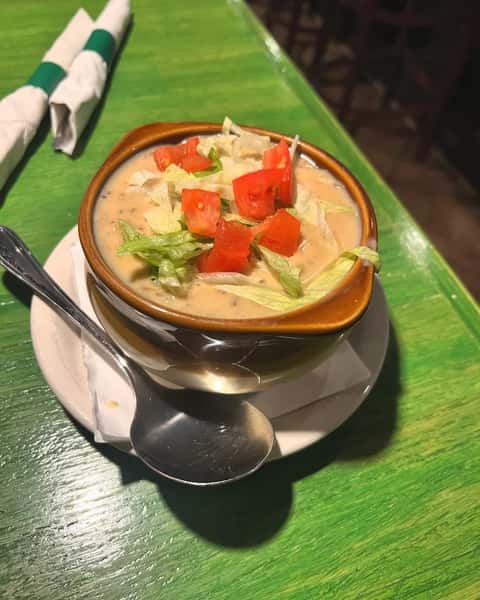 Cheesy soup topped with shredded lettuce and diced tomatoes served in a brown ceramic bowl with a spoon on the side.
