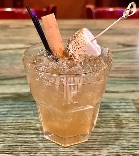 Cocktail with crushed ice, topped with a toasted marshmallow, chocolate stick, and two black straws on a wood-patterned table.