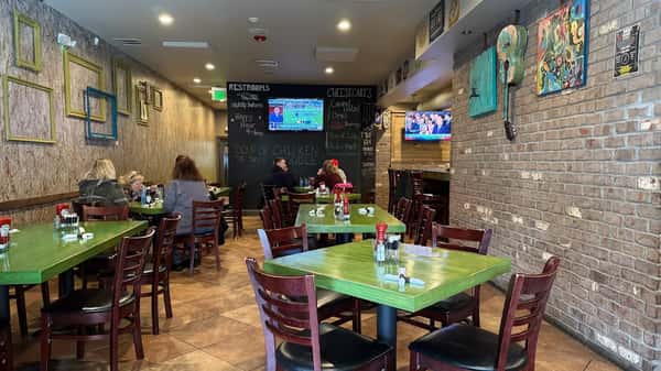 Cozy restaurant interior with green tables, wall art, flat-screen TVs, and guests dining under warm lighting.