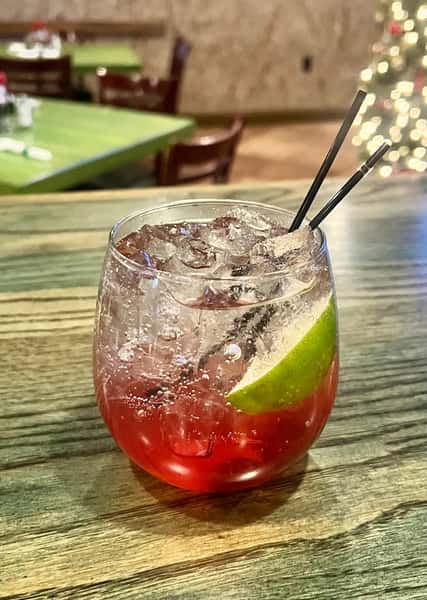Sparkling red cocktail on ice with a lime wedge and black straws, set on a wooden table with holiday lights in the background.