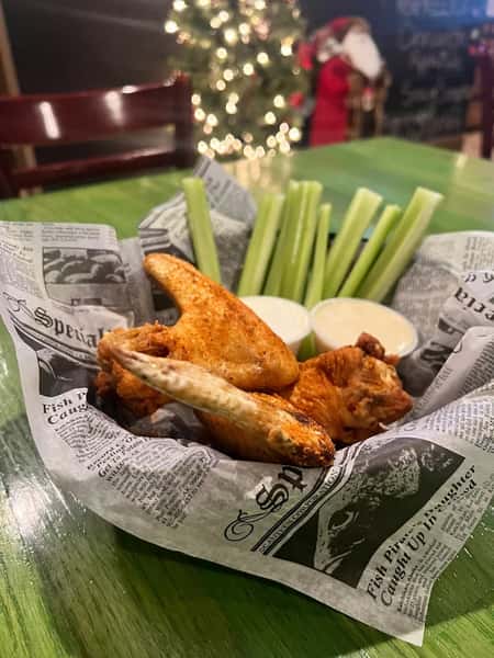 Basket of seasoned chicken wings with celery sticks and two cups of ranch dressing, served on newsprint-style paper.