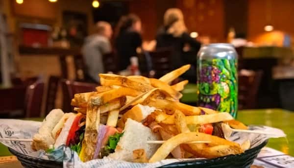 Club sandwich stacked with deli meats, lettuce, and tomato, served with crispy fries and a colorful canned drink.